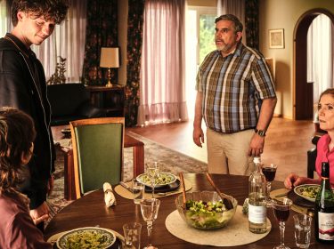 Mikes Eltern Gustav und Emilia am Tisch mit Mike und Nisha. Gustav und Mike stehen, Nisha und Emilia sitzen.