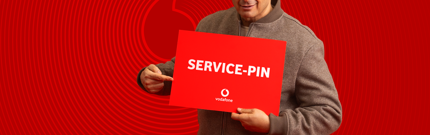 Kaya Yanar ist auf dem Bild zur Vodafone Service-PIN Kampagne zu sehen, wie er ein rotes Schild mit der Aufschrift „SERVICE-PIN“ hält und darauf zeigt.