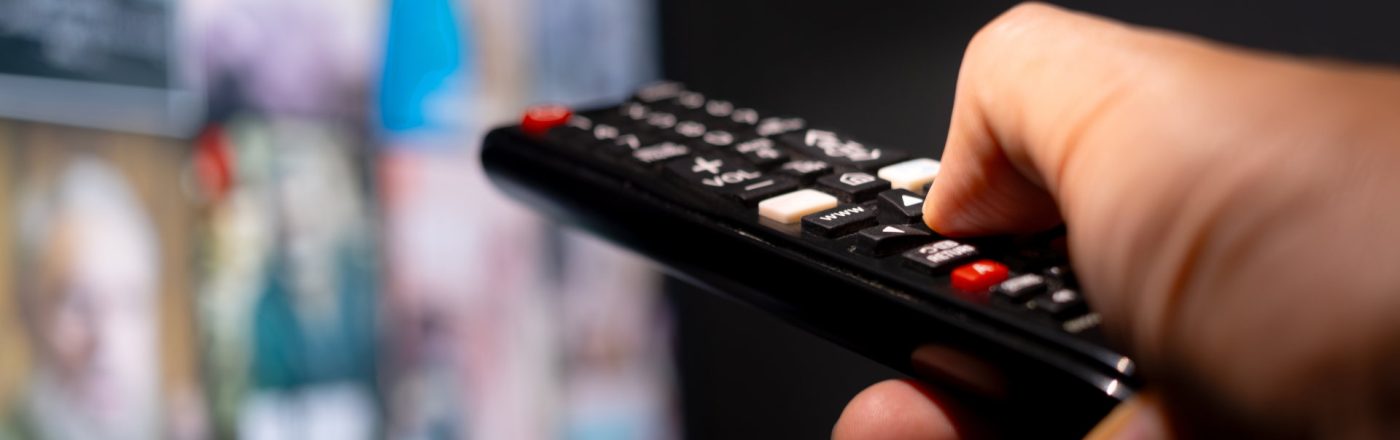 Eine Hand hält eine Fernbedienung und zeigt auf einen TV, der im verschwommen im Hintergrund zu sehen ist.