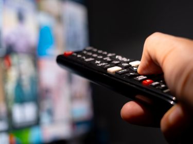 Eine Hand hält eine Fernbedienung und zeigt auf einen TV, der im verschwommen im Hintergrund zu sehen ist.