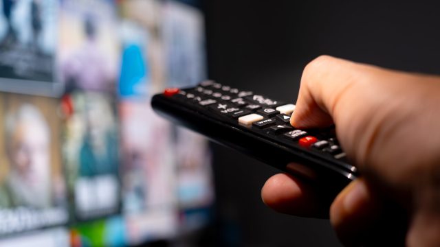 Eine Hand hält eine Fernbedienung und zeigt auf einen TV, der im verschwommen im Hintergrund zu sehen ist.