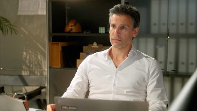 Richard (Florian Odendahl) sitz an seinem Schreibtisch und schaut nachdenklich nach oben. Vor ihm steht ein Laptop auf dem Tisch.