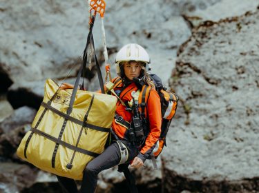 Katharina Strasser (Luise Bähr) hängt vor einer Felswand an einem Rettungstau. Ebenfalls am Tau ist ein gelber zusammengelegter Bergesack befestigt.