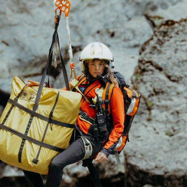 Katharina Strasser (Luise Bähr) hängt vor einer Felswand an einem Rettungstau. Ebenfalls am Tau ist ein gelber zusammengelegter Bergesack befestigt.