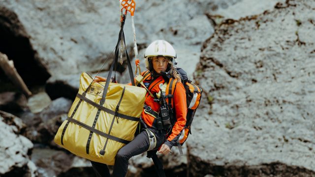 Katharina Strasser (Luise Bähr) hängt vor einer Felswand an einem Rettungstau. Ebenfalls am Tau ist ein gelber zusammengelegter Bergesack befestigt.