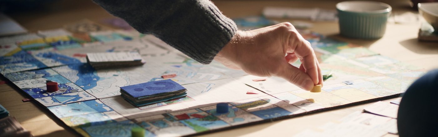 Auf dem Bild zu Brettspiel-Filme ist eine Szene am Spieltisch zu sehen, bei der eine Hand eine gelbe Spielfigur auf einem bunt gestalteten Spielbrett platziert. Rund um das Brett liegen Karten, Spielgeld und Spielsteine. Die Stimmung wirkt entspannt und familiär, Tageslicht fällt sanft auf die Szene.