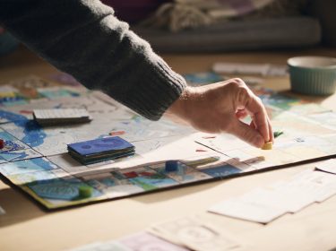 Auf dem Bild zu Brettspiel-Filme ist eine Szene am Spieltisch zu sehen, bei der eine Hand eine gelbe Spielfigur auf einem bunt gestalteten Spielbrett platziert. Rund um das Brett liegen Karten, Spielgeld und Spielsteine. Die Stimmung wirkt entspannt und familiär, Tageslicht fällt sanft auf die Szene.