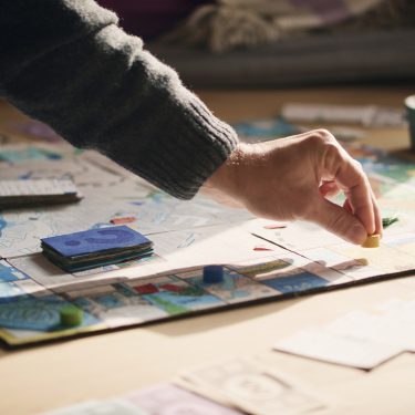 Auf dem Bild zu Brettspiel-Filme ist eine Szene am Spieltisch zu sehen, bei der eine Hand eine gelbe Spielfigur auf einem bunt gestalteten Spielbrett platziert. Rund um das Brett liegen Karten, Spielgeld und Spielsteine. Die Stimmung wirkt entspannt und familiär, Tageslicht fällt sanft auf die Szene.