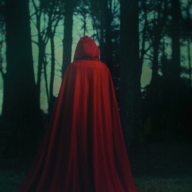 Dieses Symbolbild zur Alchemised Verfilmung zeigt Helena als abstrakte Figur in einem langen, tiefroten Umhang. Sie steht mit dem Rücken zur Kamera in einem dunklen Wald. Die Inszenierung erzeugt eine geheimnisvolle, atmosphärisch verdichtete Stimmung, die Helenass innere Reise sinnbildlich widerspiegelt.