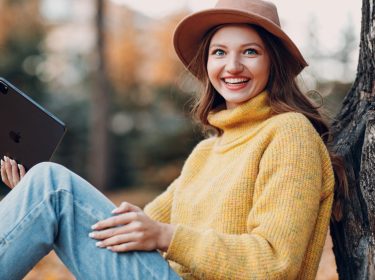 Ein junge Frau lehnt an einem Baum. Sie hält ein iPad in der Hand und lächelt in die Kamera.