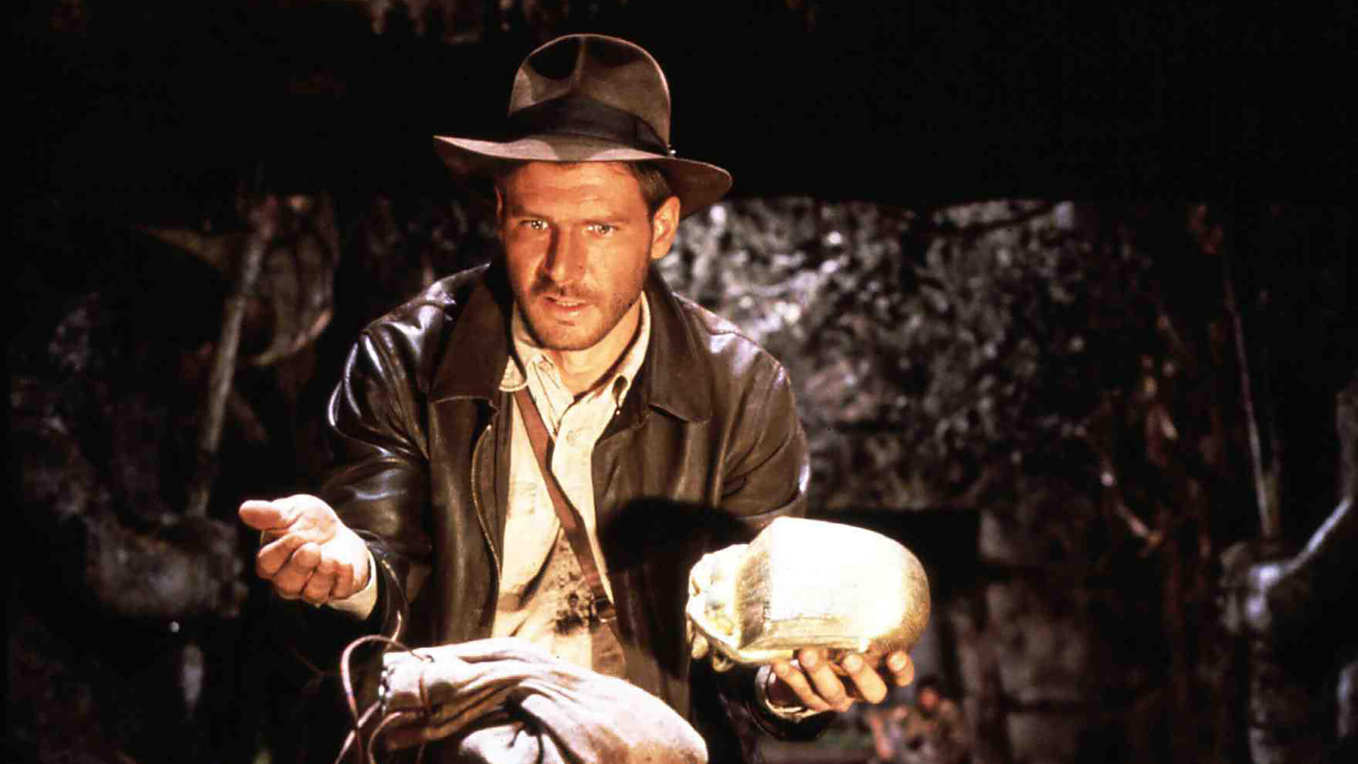Auf dem Bild aus Jäger des verlorenen Schatzes ist Harrison Ford als Indiana Jones zu sehen – einer der legendären Actionhelden der 80er und 90er. Er trägt seinen typischen Fedora-Hut und Lederjacke, während er in einer spannungsgeladenen Szene eine goldene Statue in den Händen hält. Sein Blick ist konzentriert und entschlossen.