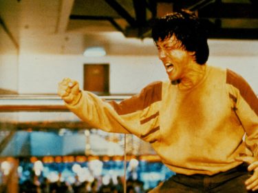 Das Bild zeigt Jackie Chan in einer ikonischen Szene aus Police Story. Schweißgebadet und voller Energie schreit er kampfbereit mit geballter Faust – typisch für seine Rolle als einer der kompromisslosesten Actionhelden der 80er und 90er. Der Hintergrund zeigt ein Einkaufszentrum, in dem gerade eine intensive Actionszene stattfindet.