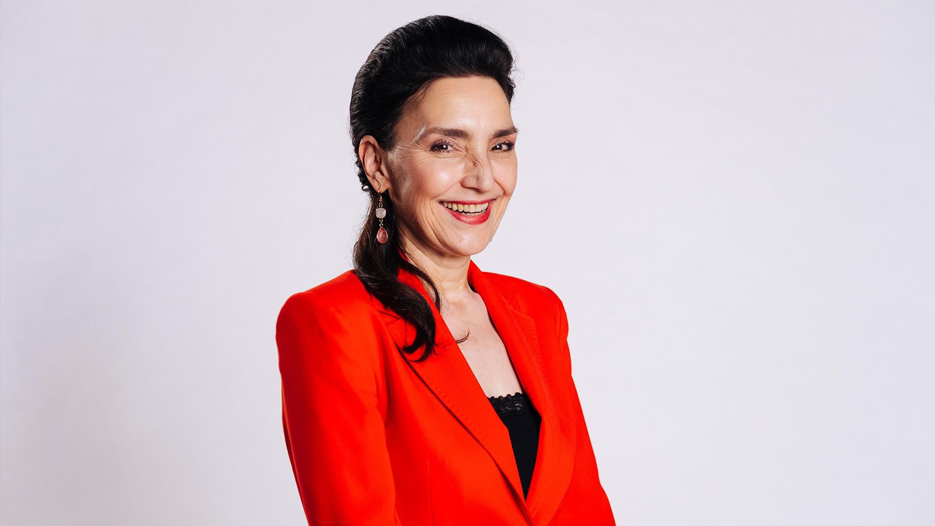 Auf diesem Bild zur Alles was zählt Besetzung ist Schauspielerin Tatjana Clasing zu sehen, die in der Serie die Rolle der Simone Steinkamp spielt. Sie trägt ein auffälliges rotes Jackett über einem schwarzen Top und lächelt herzlich in die Kamera. Ihr dunkles Haar ist elegant zurückgestylt, der Hintergrund ist hell und neutral gehalten.