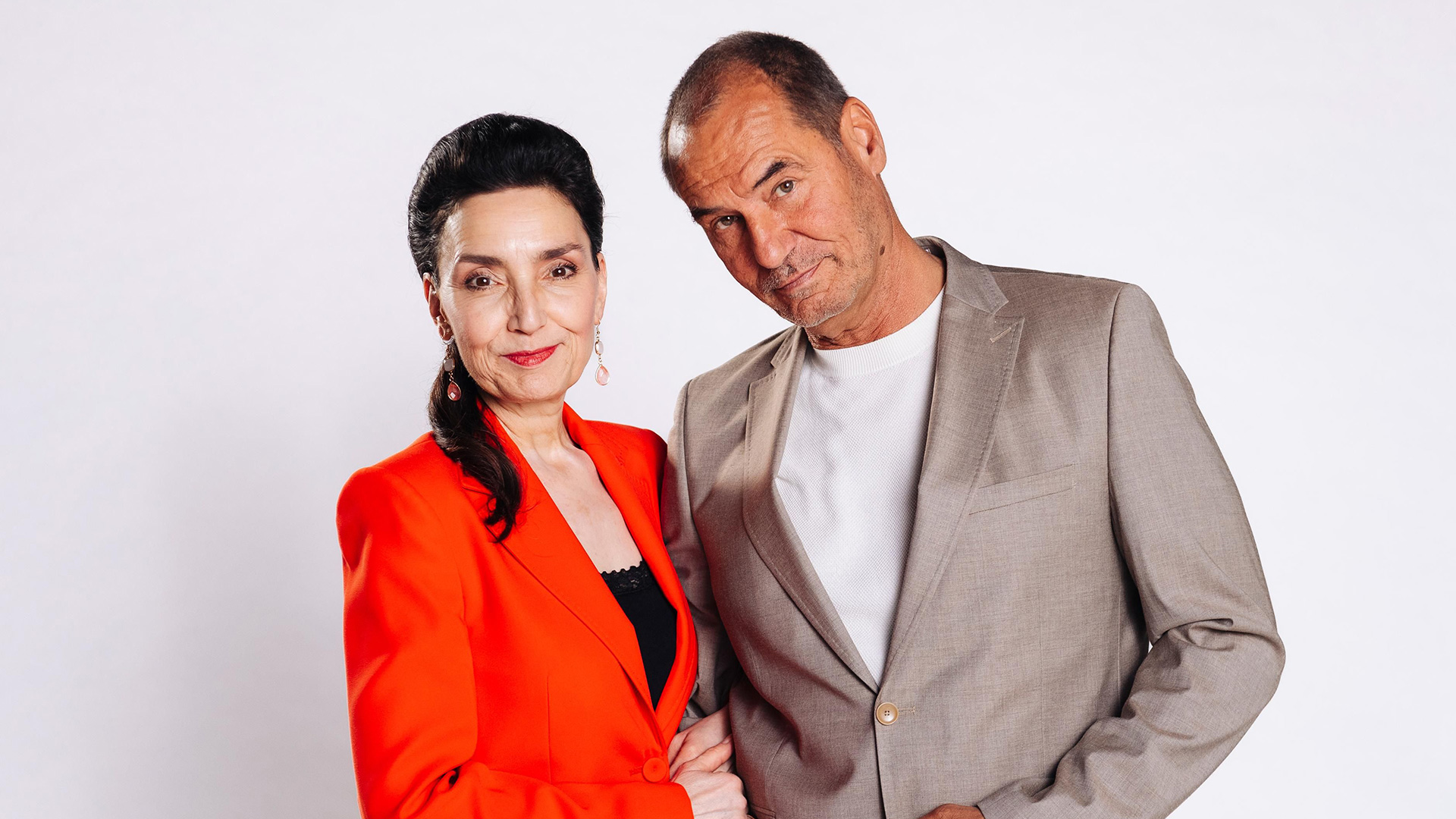 Auf dem Bild zur Alles was zählt Besetzung sind links Schauspielerin Tatjana Clasing und rechts Silvan-Pierre Leirich zu sehen. Tatjana Clasing trägt ein auffälliges rotes Jackett und lächelt dezent. Silvan-Pierre Leirich steht im beigefarbenen Anzug neben ihr und blickt entspannt in die Kamera. Der Hintergrund ist schlicht weiß.