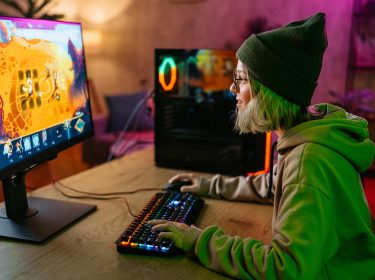 Beim Cloud-Gaming Vergleich zeigt dieses Bild eine junge Person mit grüner Mütze und Kapuzenpullover, die an einem RGB-beleuchteten Gaming-Setup spielt, während ein farbenfrohes Spiel auf dem Monitor läuft.