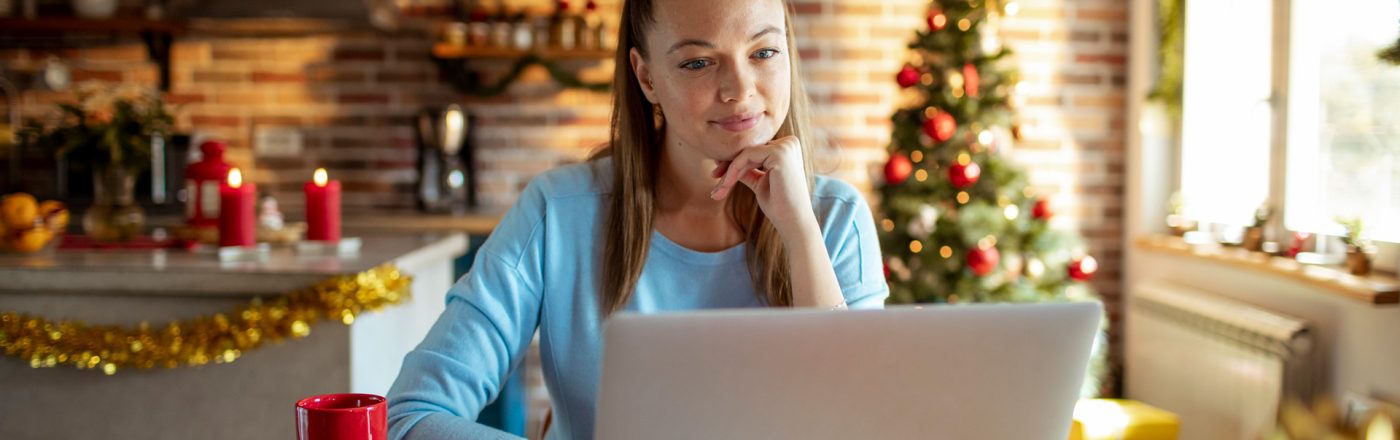 Eine junge Frau sitzt in weihnachtlich dekorierter Küche vor einem Laptop und wirkt konzentriert. Im Hintergrund steht ein geschmückter Weihnachtsbaum, Lichterketten und Kerzen sorgen für gemütliche Stimmung. Auf dem Tisch steht eine rote Tasse. Das Bild symbolisiert kreatives Arbeiten in der Adventszeit und passt zum Thema KI Prompts zu Weihnachten.