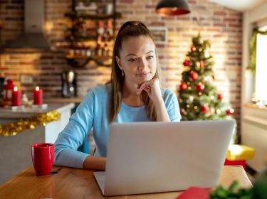 Eine junge Frau sitzt in weihnachtlich dekorierter Küche vor einem Laptop und wirkt konzentriert. Im Hintergrund steht ein geschmückter Weihnachtsbaum, Lichterketten und Kerzen sorgen für gemütliche Stimmung. Auf dem Tisch steht eine rote Tasse. Das Bild symbolisiert kreatives Arbeiten in der Adventszeit und passt zum Thema KI Prompts zu Weihnachten.