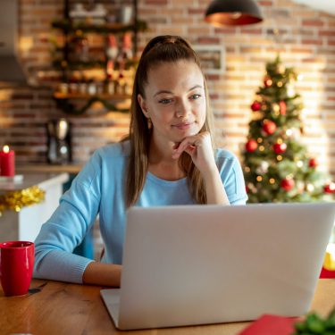 Eine junge Frau sitzt in weihnachtlich dekorierter Küche vor einem Laptop und wirkt konzentriert. Im Hintergrund steht ein geschmückter Weihnachtsbaum, Lichterketten und Kerzen sorgen für gemütliche Stimmung. Auf dem Tisch steht eine rote Tasse. Das Bild symbolisiert kreatives Arbeiten in der Adventszeit und passt zum Thema KI Prompts zu Weihnachten.