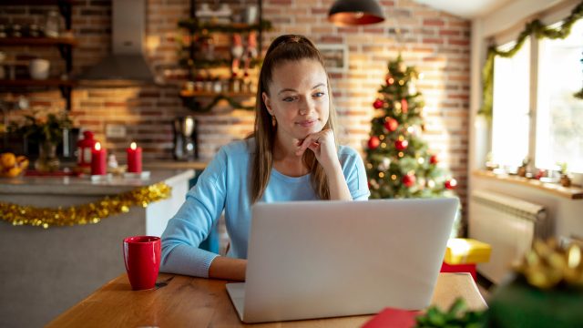 Eine junge Frau sitzt in weihnachtlich dekorierter Küche vor einem Laptop und wirkt konzentriert. Im Hintergrund steht ein geschmückter Weihnachtsbaum, Lichterketten und Kerzen sorgen für gemütliche Stimmung. Auf dem Tisch steht eine rote Tasse. Das Bild symbolisiert kreatives Arbeiten in der Adventszeit und passt zum Thema KI Prompts zu Weihnachten.