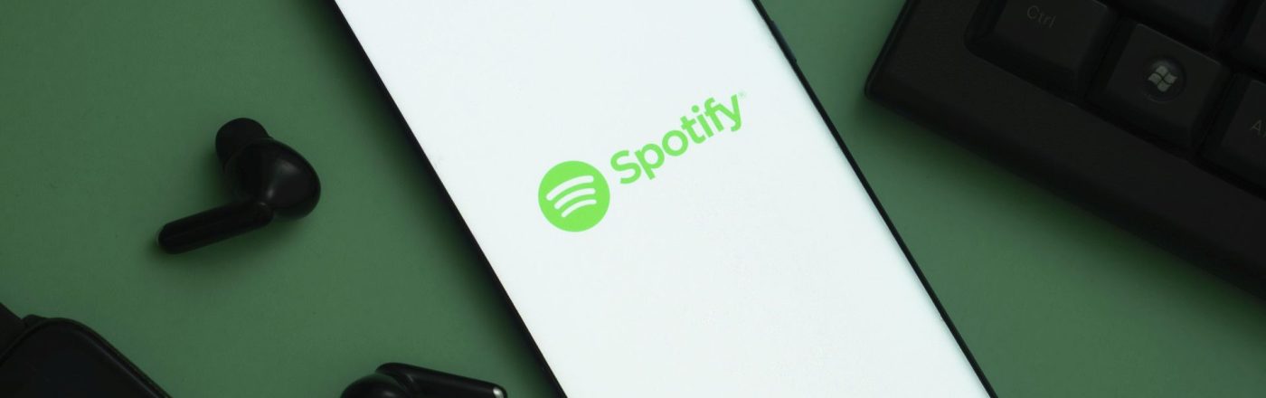 Das Logo von Spotify ist auf einem Handy zu sehen, links davon liegen schwarze Kopfhörer.