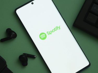 Das Logo von Spotify ist auf einem Handy zu sehen, links davon liegen schwarze Kopfhörer.