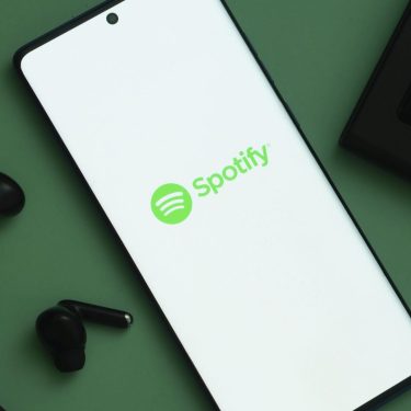 Das Logo von Spotify ist auf einem Handy zu sehen, links davon liegen schwarze Kopfhörer.