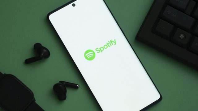 Das Logo von Spotify ist auf einem Handy zu sehen, links davon liegen schwarze Kopfhörer.