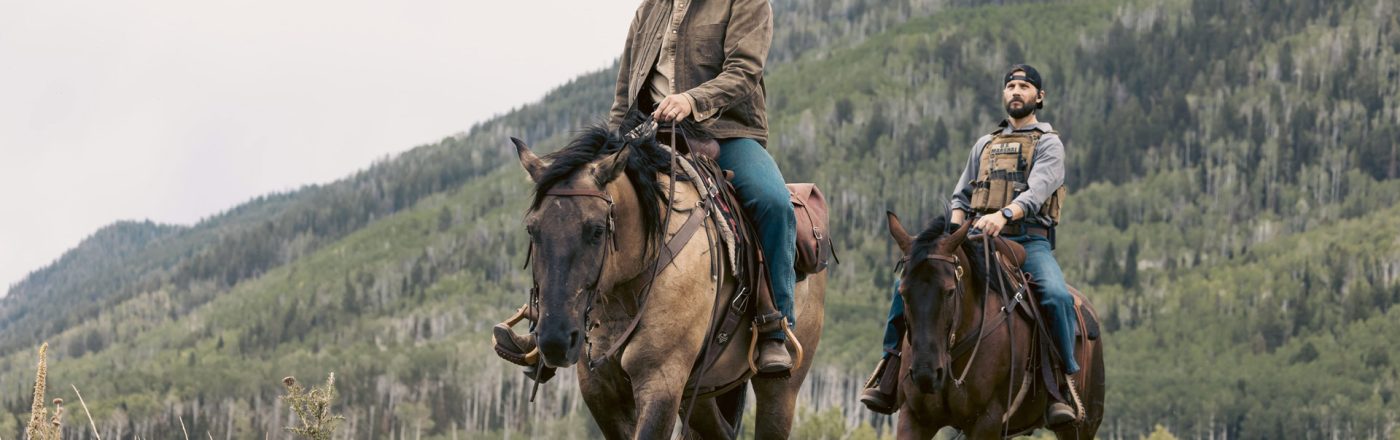 In dieser Szene aus der Marshals Serie sind Luke Grimes als Kayce Dutton und Logan Marshall-Green als Pete Calvin auf Pferden in einer bergigen, waldreichen Landschaft unterwegs. Beide tragen lässige Westernkleidung mit ernsten Gesichtsausdrücken. Die Stimmung wirkt ruhig, aber gespannt – typisch für ein modernes Western-Drama.