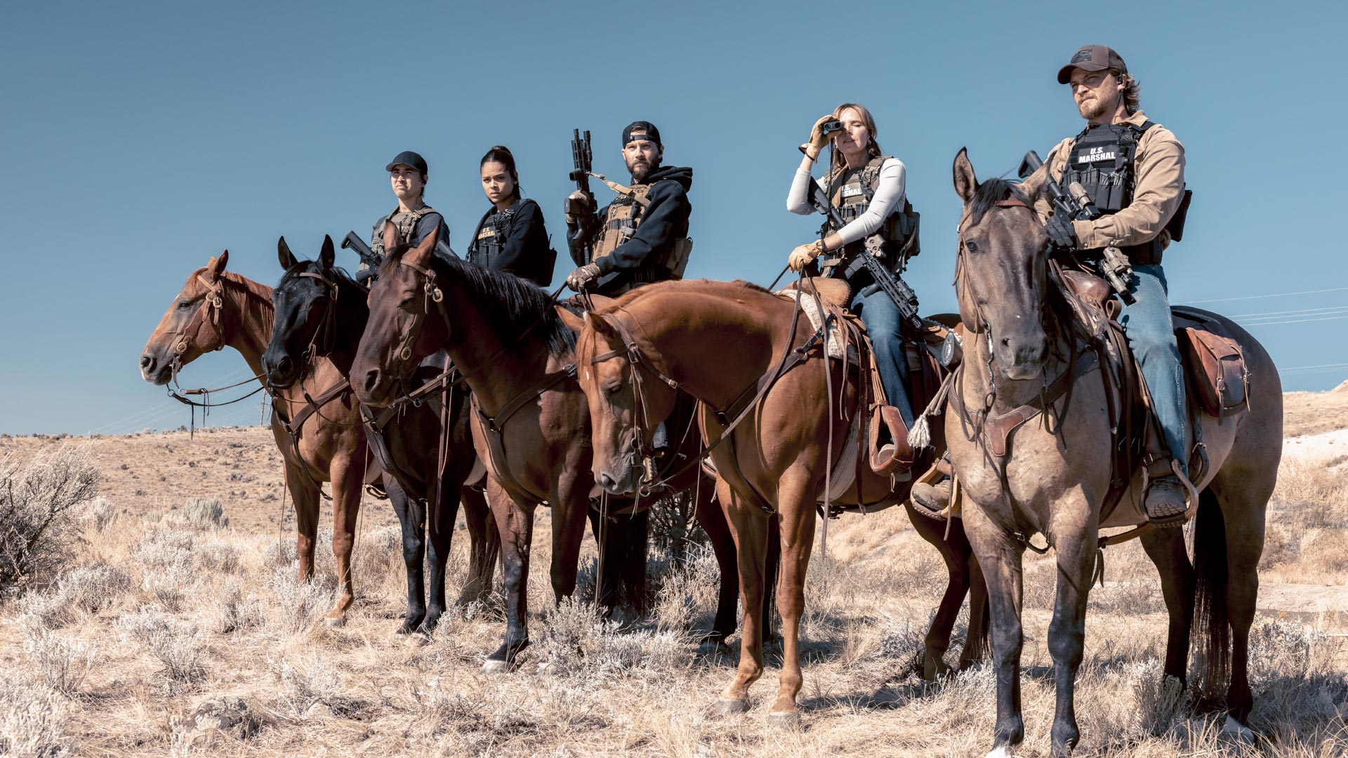 In dieser Szene aus der Marshals Serie sind fünf U.S. Marshals hoch zu Pferd in einer trockenen, kargen Landschaft zu sehen. Von links nach rechts: Ash Santos, Tatanka Means, Logan Marshall-Green als Pete Calvin, Arielle Kebbel als Belle mit Fernglas, und Luke Grimes als Kayce Dutton. Alle tragen kugelsichere Westen und wirken entschlossen und einsatzbereit.