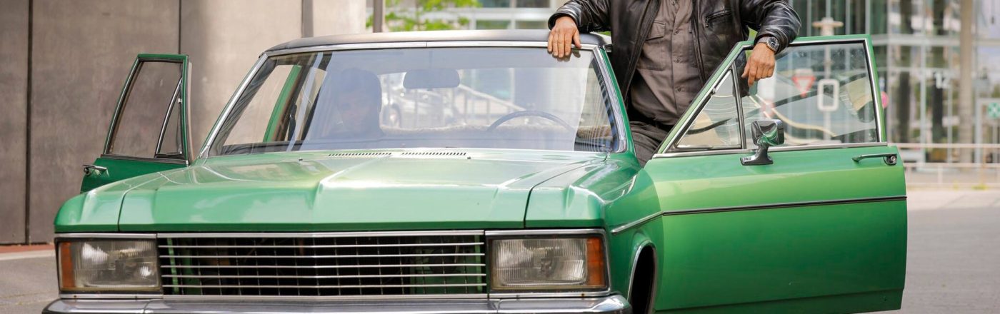 Auf dem Bild in den GigaTV-Highlights im Dezember 2025 siehst du eine Szene aus „Der letzte Bulle“: Mick Brisgau (Henning Baum) steht lässig an einem grünen Oldtimer mit offener Tür in einer urbanen Umgebung.