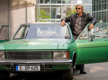 Auf dem Bild in den GigaTV-Highlights im Dezember 2025 siehst du eine Szene aus „Der letzte Bulle“: Mick Brisgau (Henning Baum) steht lässig an einem grünen Oldtimer mit offener Tür in einer urbanen Umgebung.