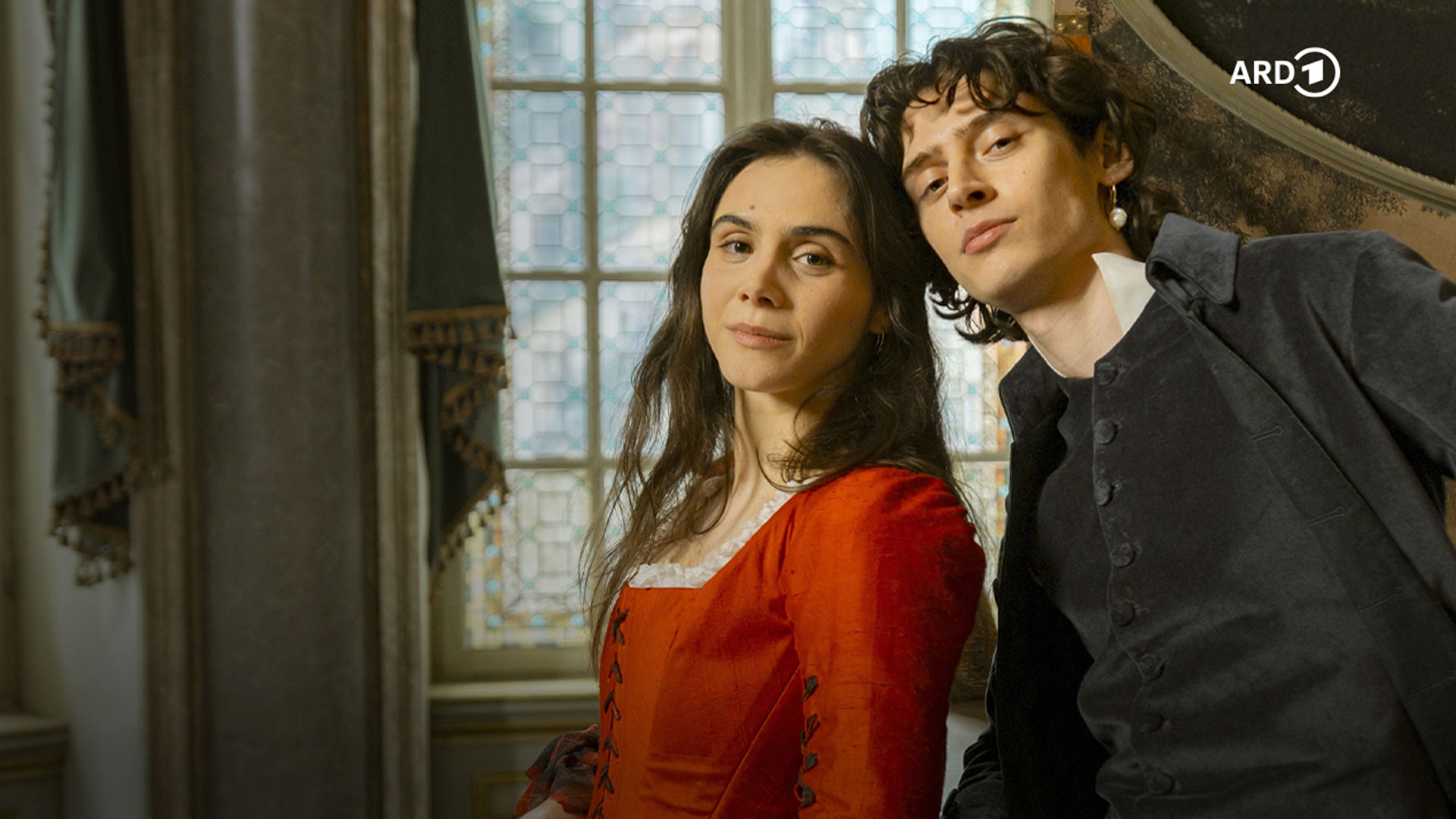 Auf dem Bild in den GigaTV-Highlights im Dezember 2025 siehst du eine Szene aus „Mozart/Mozart“: Wolfgang Amadeus Mozart (Eren M. Güvercin) und seine Schwester Nannerl (Havana Joy) posieren in historischer Kleidung vor einem Fenster mit Bleiglas.