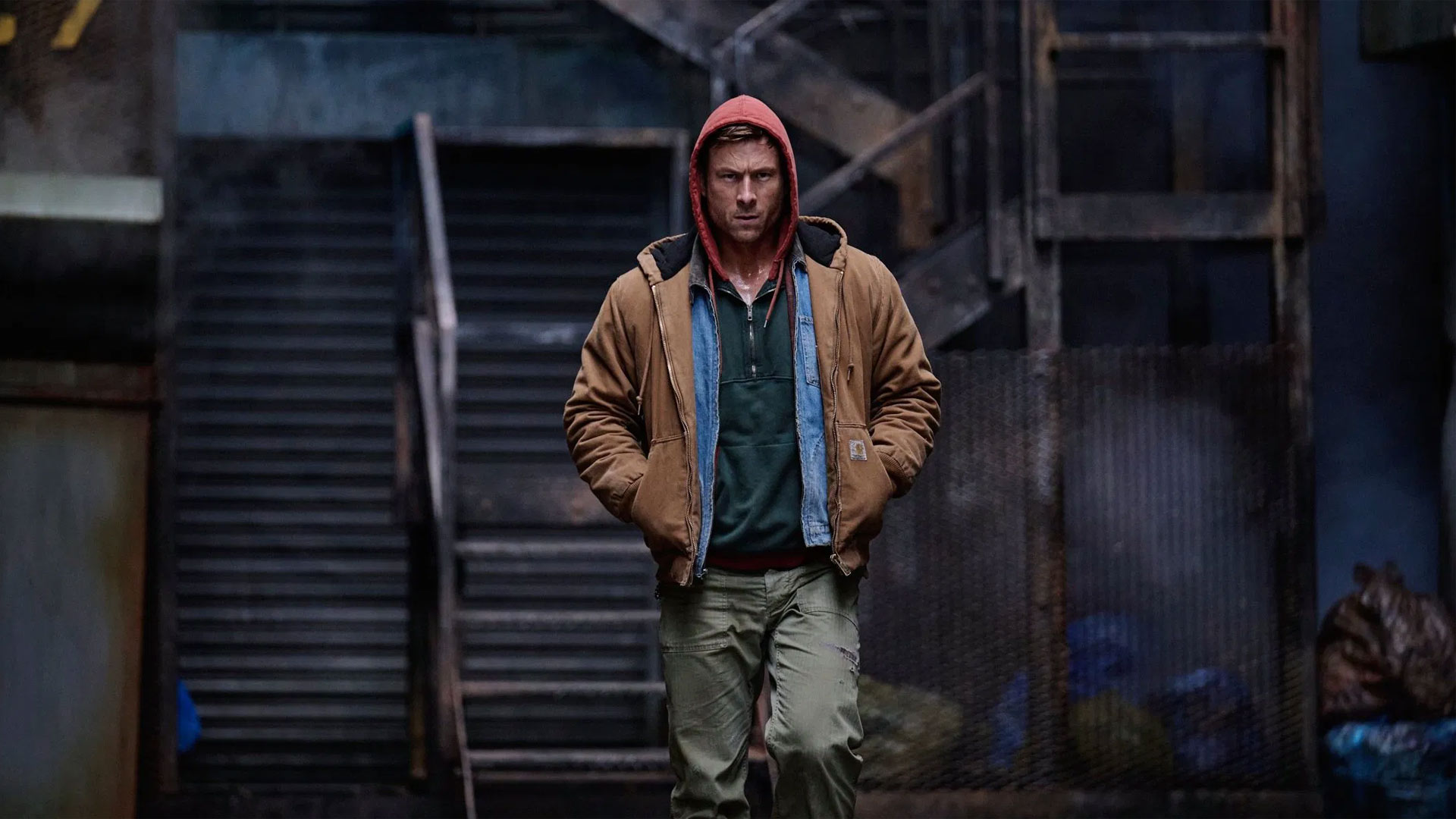 Auf dem Bild in den GigaTV-Highlights im Dezember 2025 siehst du eine Szene aus „The Running Man“: Ben Richards (Glen Powell) geht entschlossen durch eine düstere Gasse mit industriellem Hintergrund, bekleidet mit Kapuzenpulli und Jacke.