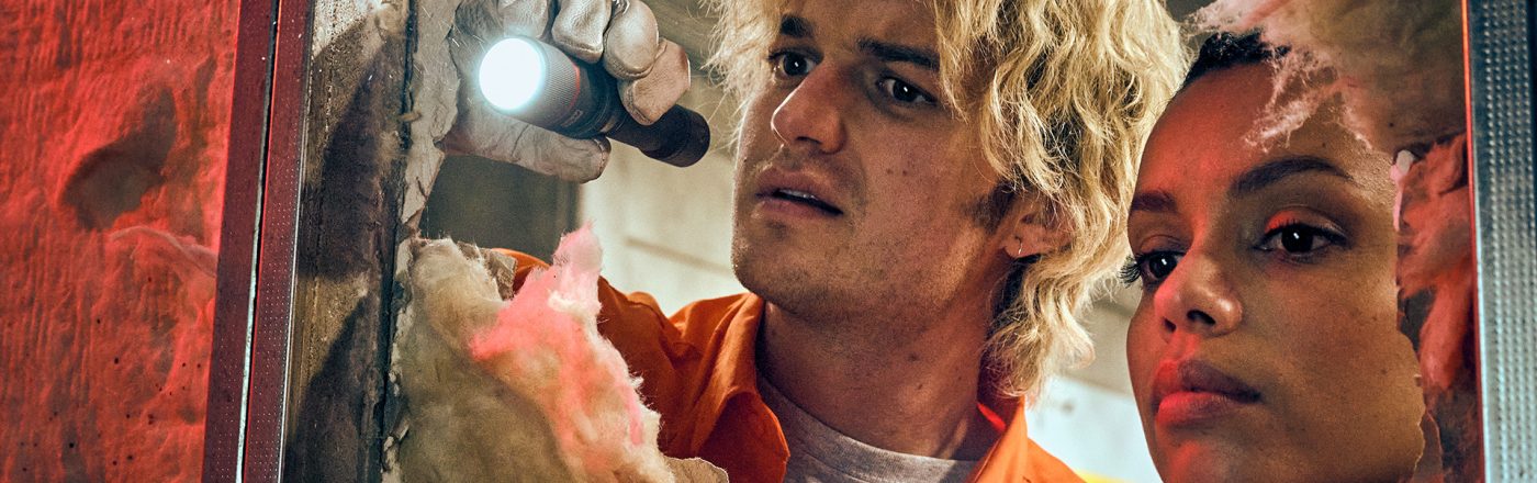 Auf dem Bild aus dem Film Cold Storage sind die Charaktere Teacake, gespielt von Joey Keery, und Naomi, dargestellt von Georgina Campbell, bei einer spannenden Untersuchungsszene zu sehen. Beide schauen konzentriert durch ein Loch in einer Wand und leuchten mit Taschenlampen in die Dunkelheit. Die Atmosphäre wirkt angespannt und geheimnisvoll.