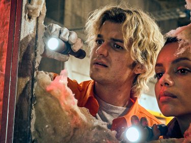 Auf dem Bild aus dem Film Cold Storage sind die Charaktere Teacake, gespielt von Joey Keery, und Naomi, dargestellt von Georgina Campbell, bei einer spannenden Untersuchungsszene zu sehen. Beide schauen konzentriert durch ein Loch in einer Wand und leuchten mit Taschenlampen in die Dunkelheit. Die Atmosphäre wirkt angespannt und geheimnisvoll.