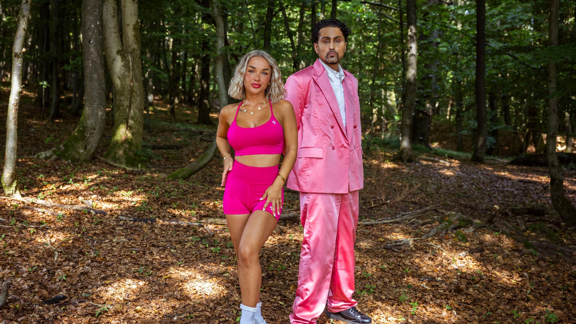 Das Bild aus der Couple Challenge 2026 zeigt Emmy und Samuel im Wald. Emmy trägt ein sportliches pinkes Outfit mit Shorts und Top, während Samuel in einem auffälligen glänzenden rosa Anzug mit weißem Hemd danebensteht. Beide schauen selbstbewusst in die Kamera und setzen auf einen stilvollen und starken Auftritt.