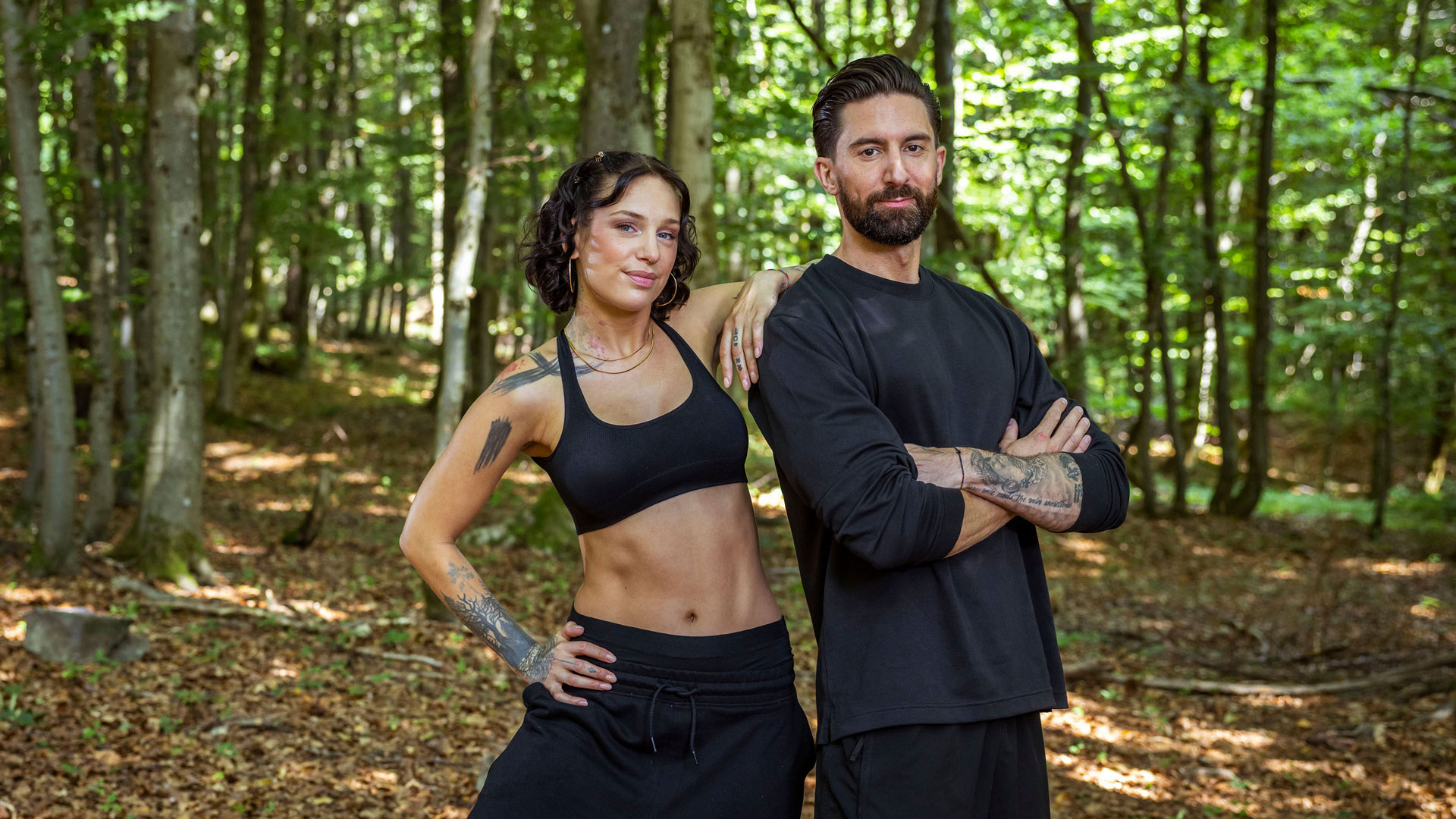 Auf dem Bild aus der Couple Challenge 2026 stehen Liza und Sebi selbstbewusst im Wald. Beide tragen schwarze Sportkleidung – Liza ein Sport-Bralette und Sebi ein langärmliges Shirt. Liza legt locker den Arm auf Sebis Schulter, während er mit verschränkten Armen in die Kamera blickt. Ihre Ausstrahlung wirkt ruhig, fokussiert und entschlossen.