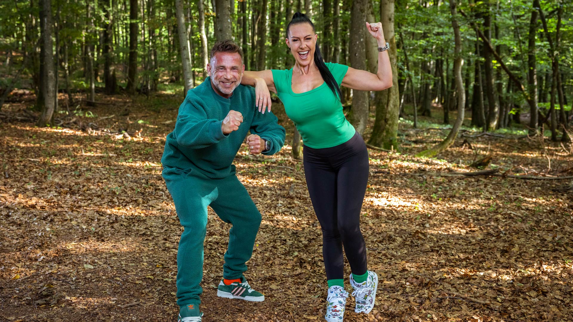 Auf dem Bild aus der Couple Challenge 2026 sind Patti und Patrick in sportlicher Siegerpose im Wald zu sehen. Patrick trägt einen grünen Fleece-Trainingsanzug, Patti ein grünes T-Shirt mit schwarzer Leggings. Beide lachen breit, zeigen starke Posen mit geballten Fäusten und strahlen pure Motivation und Wettkampfgeist aus.