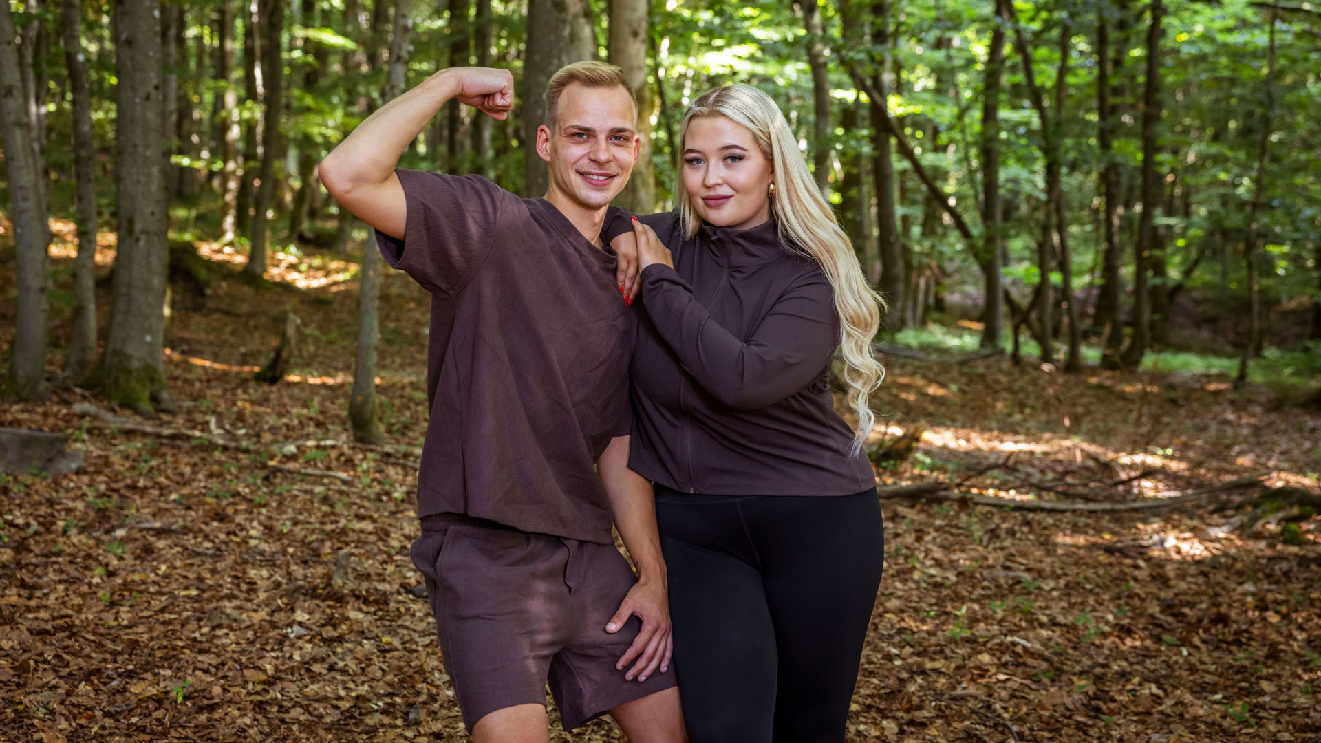 Auf dem Bild aus der Couple Challenge 2026 stehen Sophie und Hendrik in brauner Sportkleidung im Wald. Hendrik zeigt mit einem Lächeln seinen Bizeps, während Sophie sich entspannt an ihn anlehnt und selbstbewusst in die Kamera blickt. Beide wirken harmonisch und bereit für die Herausforderungen des Wettbewerbs.