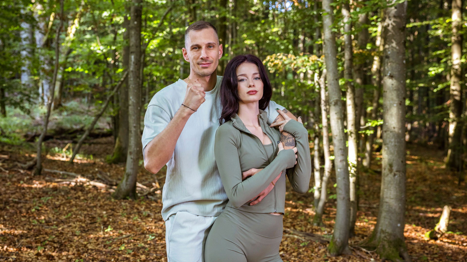 Auf dem Bild aus der Couple Challenge 2026 posieren Zoe und Patrik ernst und fokussiert im Wald. Zoe trägt ein eng anliegendes, olivgrünes Sportoutfit mit langen Ärmeln, Patrik ein helles Shirt mit passender Hose. Beide zeigen eine kämpferische Haltung – Patrik mit geballter Faust, Zoe mit verschränkten Armen und intensivem Blick.
