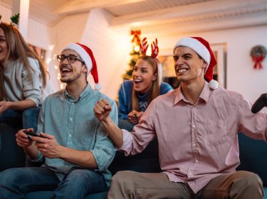 Auf dem Bild in dem Artikel zu den Spielen zu Weihnachten sind vier junge Erwachsene zu sehen, die auf einem Sofa sitzen und gemeinsam Videospiele spielen.