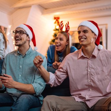 Auf dem Bild in dem Artikel zu den Spielen zu Weihnachten sind vier junge Erwachsene zu sehen, die auf einem Sofa sitzen und gemeinsam Videospiele spielen.