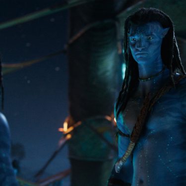 Auf dem Bild ist Jake Sully, gespielt von Sam Worthington, in einer Szene aus dem Film Avatar: The Way of Water zu sehen. Er steht im Vordergrund in traditioneller Kleidung der Na'vi und blickt ernst nach vorn. Die nächtliche Kulisse mit bläulichem Licht verstärkt die mystische Atmosphäre. Das Bild eignet sich perfekt für Inhalte rund um Avatar-Fakten.