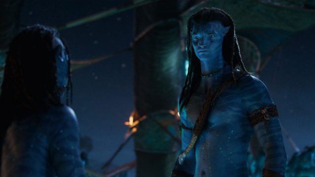 Auf dem Bild ist Jake Sully, gespielt von Sam Worthington, in einer Szene aus dem Film Avatar: The Way of Water zu sehen. Er steht im Vordergrund in traditioneller Kleidung der Na'vi und blickt ernst nach vorn. Die nächtliche Kulisse mit bläulichem Licht verstärkt die mystische Atmosphäre. Das Bild eignet sich perfekt für Inhalte rund um Avatar-Fakten.