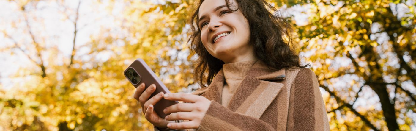 Lächelnde Frau mit Mantel steht im herbstlichen Park und hält mit beiden Händen ein iPhone