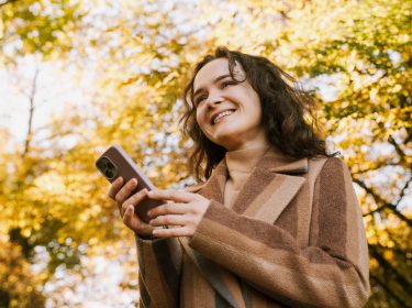 Lächelnde Frau mit Mantel steht im herbstlichen Park und hält mit beiden Händen ein iPhone
