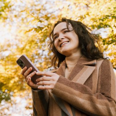 Lächelnde Frau mit Mantel steht im herbstlichen Park und hält mit beiden Händen ein iPhone