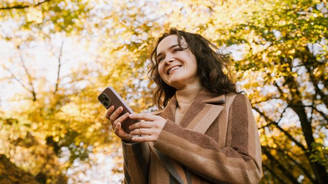 Lächelnde Frau mit Mantel steht im herbstlichen Park und hält mit beiden Händen ein iPhone
