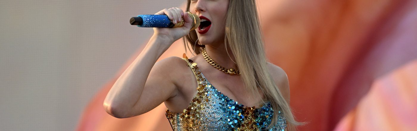Das Bild zeigt Taylor Swift bei einem Live-Auftritt und dient symbolisch für Artikel über Taylor Swift-Dokus. Sie trägt ein glitzerndes, gold-blaues Bühnenoutfit und singt mit ausdrucksstarkem Gesichtsausdruck in ein blaues Mikrofon. Der verschwommene Hintergrund unterstreicht die emotionale und kraftvolle Atmosphäre der Szene.