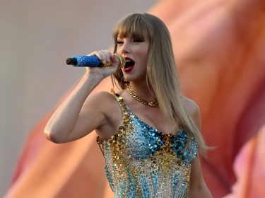 Das Bild zeigt Taylor Swift bei einem Live-Auftritt und dient symbolisch für Artikel über Taylor Swift-Dokus. Sie trägt ein glitzerndes, gold-blaues Bühnenoutfit und singt mit ausdrucksstarkem Gesichtsausdruck in ein blaues Mikrofon. Der verschwommene Hintergrund unterstreicht die emotionale und kraftvolle Atmosphäre der Szene.